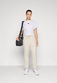 Biela kratká tričko s krátkymi rukávmi a čiernym logom, béžové zúžené jogger nohavice a biele tenisky. Čierna taška cez rameno so strapcom a textúrovaným povrchom.