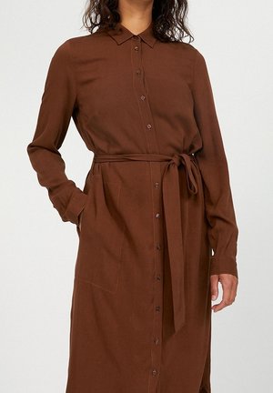 Femme portant une robe boutonnée marron à manches longues avec une ceinture nouée à la taille et des poches latérales, sur un fond uni.