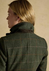 Vihreä villatakki, jossa on oransseja ja purppuran värisiä ruutukuoseja. Varustettu ruskealla mokkasella kauluksella, jossa on kohokuvioitu logo ja kultaiset varusteet.