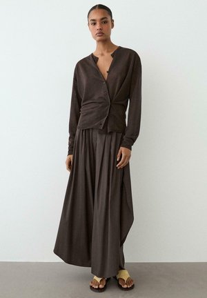 Donna in piedi contro una parete bianca che indossa un cardigan marrone scuro con bottoni, gonna a gamba larga abbinata e sandali beige con anello per le dita.