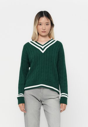 Ung kvinne med rett hår iført en grønn kabelstrikket genser med hvite striper og lys grå bukser, står mot en hvit bakgrunn.