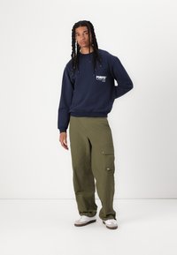 Marinegenser med "POMPEII" grafikk, olivengrønne cargo-bukser med lommedetaljer, og hvite sneakers med brune detaljer.