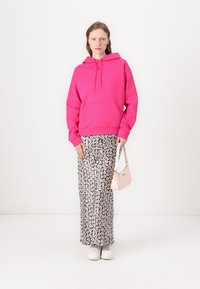 Felpa rosa brillante con tasca frontale, abbinata a una gonna maxi floreale in bianco e nero. Il modello tiene una piccola borsa rosa. Sneakers bianche.