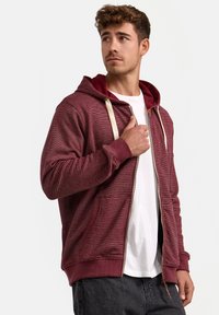 Bordeauxfarbener Reißverschluss-Hoodie aus strukturiertem Stoff mit cremefarbenen Kordeln, Kängurutaschen und gerippten Bündchen. Getragen über einem weißen T-Shirt.