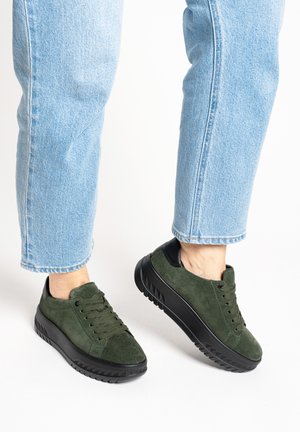 Person wearing light blue jeans and dark green suede platform sneakers with black soles against a white background.