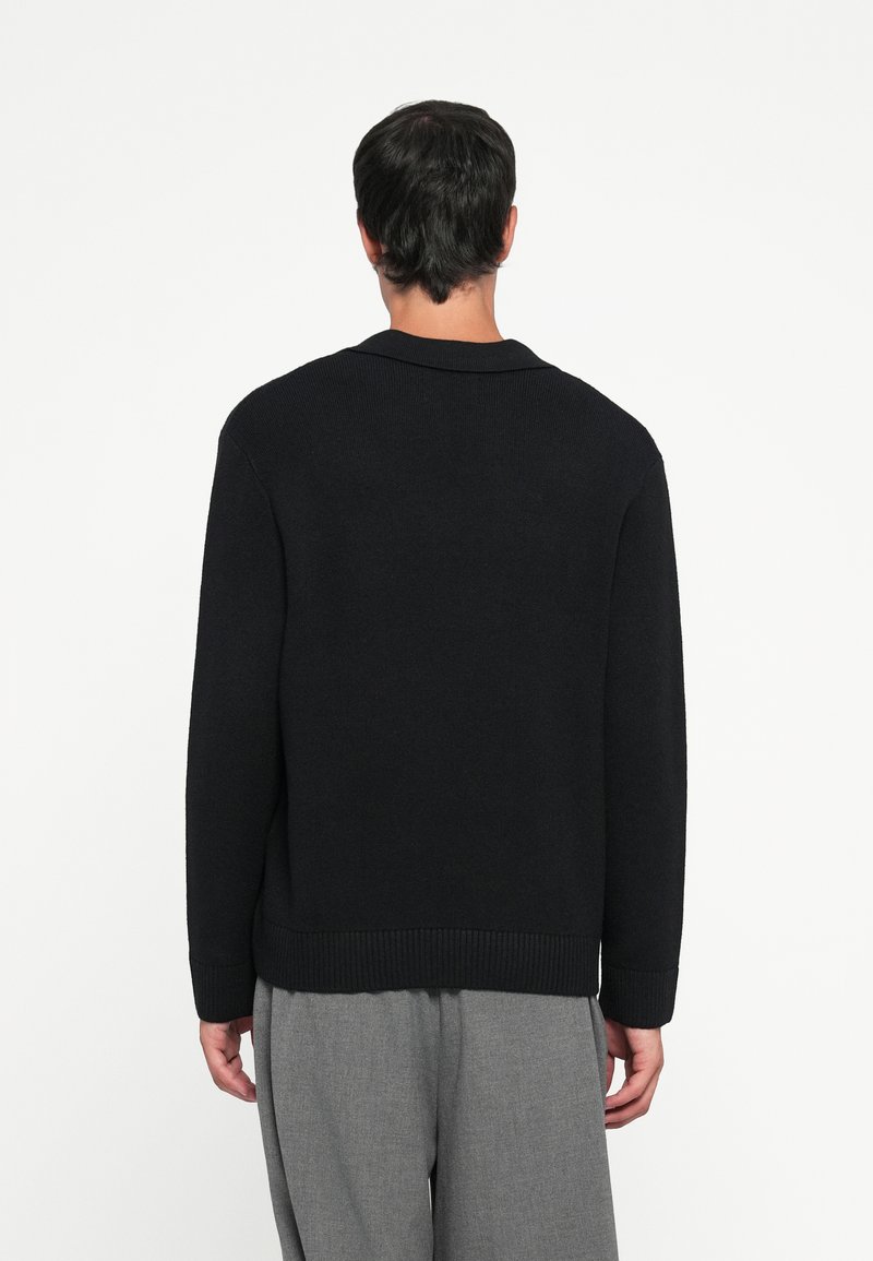 Maglione nero lavorato a maglia con orlo e colletto a costine, dotato di maniche lunghe e vestibilità morbida. Indossato sopra pantaloni grigi chiari.