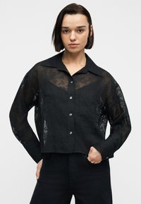 Νεαρή γυναίκα με κοντά σκούρα μαλλιά, φορώντας διαφανές μαύρο πουκάμισο με λουλουδάτο σχέδιο και μαύρο παντελόνι, μπροστά σε απλό φόντο.