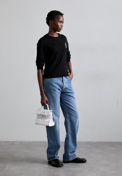 Pulover negru cu pliuri, blugi din denim bleu deschis și o geantă mică albă. Pantofi negri lustruiți completează ținuta. Modelul stă pe un fundal gri.