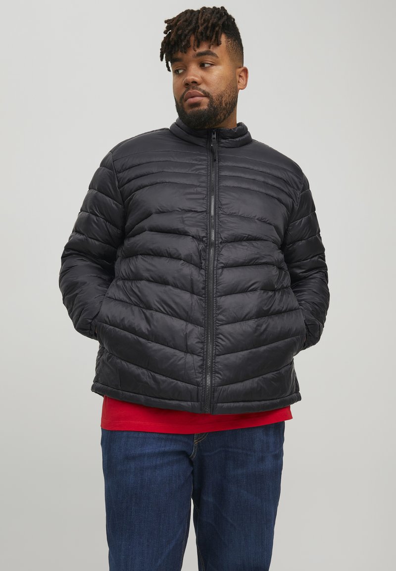 Giacca a vento nera con collo alto, motivo a righe orizzontali, zip frontale e tasche laterali, indossata sopra una maglietta rossa e jeans blu.