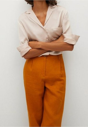 Personne debout, bras croisés, portant une chemise beige clair boutonnée avec les manches retroussées et un pantalon taille haute orange brûlé.