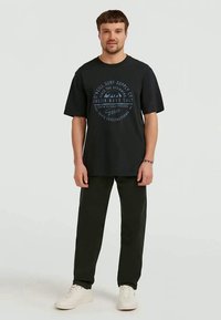 Man in een zwart graphic t-shirt, donkere broek, witte sneakers, staande tegen een effen achtergrond, recht vooruit kijkend, met ontspannen armen naast zich.