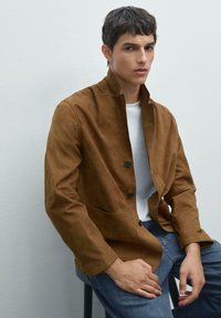 Braune Wildlederjacke mit Stehkragen, Vorderknöpfen und zwei Seitentaschen, getragen über einem weißen Hemd und blauen Jeans. Glatte Textur.