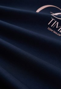 Sötétkék szövet sima textúrával, finom fényű. Tartalmaz egy világosbarna logo szöveget "TOM TAILOR", modern betűtípussal.