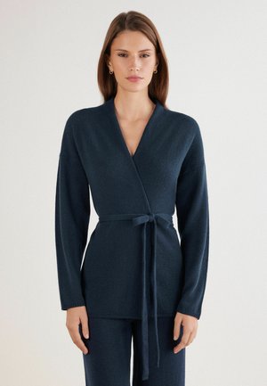Pull-over en style wrap de couleur bleu marine profond. Tissu en tricot doux, manches longues, décolleté en V et lien ajustable à la taille. Détails simples à l'ourlet.