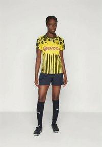 Κίτρινη φανέλα ποδοσφαίρου με γραφικό σχέδιο και μαύρες ρίγες, που φέρει το λογότυπο "Evonik", συνδυασμένη με μαύρη βερμούδα και κάλτσες μέχρι το γόνατο.