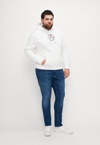 Biała bluza z kapturem z kieszenią z przodu i czarnym logo, w połączeniu z niebieskimi dżinsami. Model nosi białe sneakersy, stoi na neutralnym tle.