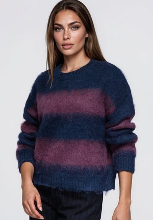 Une femme porte un pull ample et duveteux bleu marine à larges rayures bordeaux, sur un fond uni et clair.