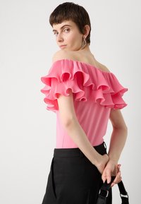 Top rosa de hombros al descubierto con pliegues con volantes, corsé ajustado y textura suave. Combinado con pantalones negros de talle alto.