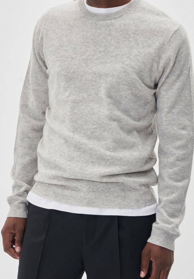 Un pull en maille gris clair avec une texture douce, présentant un col rond et des poignet côtelés, superposé sur une chemise blanche à ourlet droit.