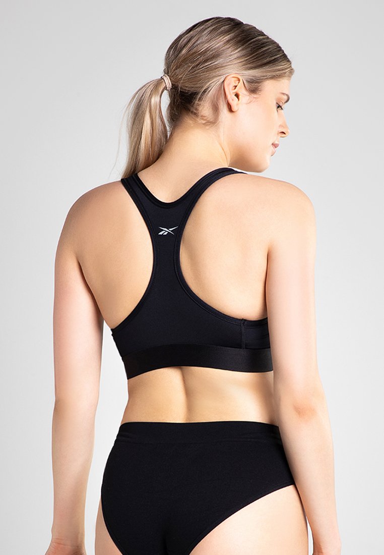 Soutien-gorge de sport noir avec un design dos nageur, fabriqué en tissu extensible. Texture lisse et branding minimal à l'arrière. Associé à des slips noirs.