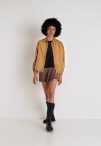 Giacca marrone con texture sopra una maglietta nera, abbinata a una gonna a righe e stivali neri fino al ginocchio. La modella sta in piedi su uno sfondo neutro.