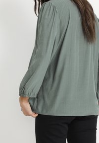 Blouse verte à manches longues avec une finition texturée, présentant une coupe décontractée, des poignets froncés et un motif rayé subtil.