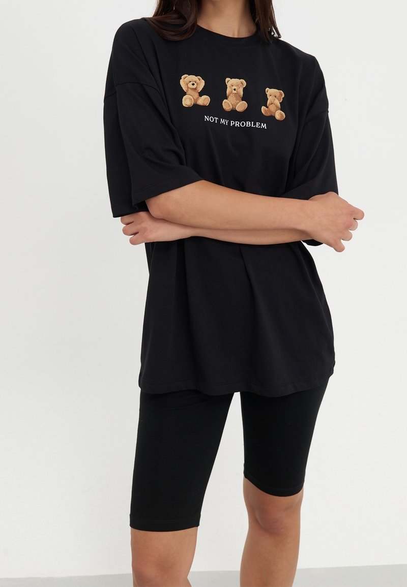 Personne portant un t-shirt noir oversize avec trois oursons en peluche et le texte "NOT MY PROBLEM" ainsi que des shorts de cycliste noirs, se tenant les bras croisés.