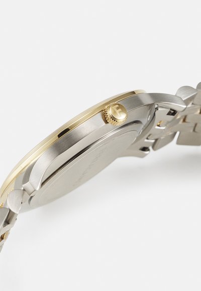 Silver and gold-tone metal wristwatch with a round case, textured bezel, and a prominent crown featuring a logo, linked metal bracelet.