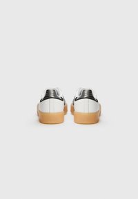 Zapatillas blancas con parte superior de cuero suave, detalles en negro y una suela de goma marrón. Presentan un diseño de perfil bajo y un talón texturizado.