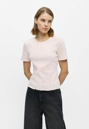 Jeune femme aux cheveux mi-longs portant un top côtelé rose clair à manches courtes et un jean noir ample, debout sur fond blanc.