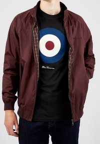 Chaqueta bomber ligera de color burdeos, con cremallera y forro de cuadros. Camiseta negra con un diseño circular en azul, blanco y rojo. Pantalones de mezclilla.