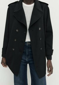 Czarny dwurzędowy płaszcz z wyciętymi klapami, o fakturowanej tkaninie i dużych guzikach. Nosi się go na prążkowany biały sweter i niebieskie dżinsy.
