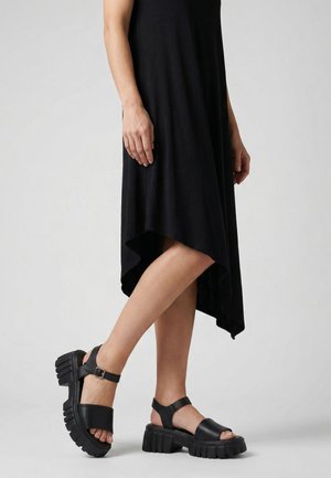 Vrouw die een zwarte asymmetrische jurk draagt en zwarte grove platformsandalen met enkelbandjes, staand tegen een grijze achtergrond.