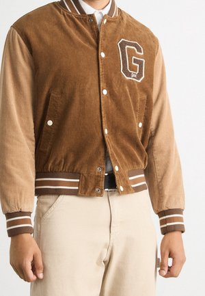 Mężczyzna noszący brązową sztruksową kurtkę varsity z naszywką z literą „G”, paskami na mankietach i kołnierzu, w połączeniu z jasnobeżowymi spodniami i czarnym paskiem.