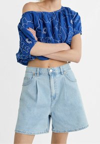 Blusa azul, sin mangas y con escote off-the-shoulder, con bordado floral blanco; combinada con pantalones cortos de mezclilla de lavado claro con pinzas y bolsillos frontales.