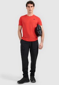 Homme debout, portant un T-shirt rouge, un pantalon cargo noir et des bottes noires, tenant une doudoune noire sous le bras.