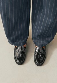 Schwarze Lackleder-Loafers mit dekorativem Quast und Kettendetail, kombiniert mit marineblauen Nadelstreifenhosen.