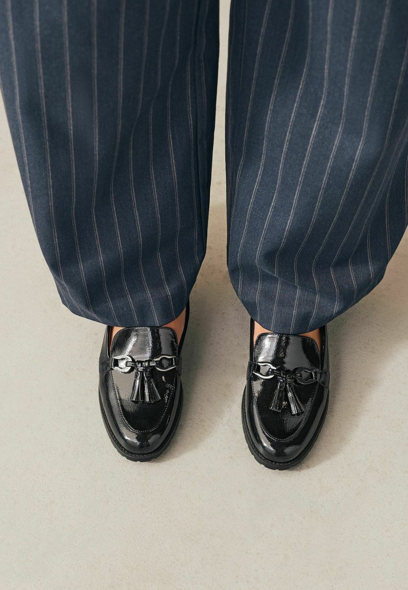 Schwarze Lackleder-Loafers mit dekorativem Quast und Kettendetail, kombiniert mit marineblauen Nadelstreifenhosen.