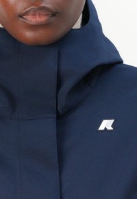 Veste coupe-vent bleu marine avec un col haut, dotée d'un patch logo blanc et d'une texture lisse. Le design est conçu pour la fonctionnalité.
