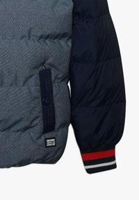 Vadderad jacka med en grå houndstooth-kropp och marinblå ärmar. Innehåller ribbade ärmslut med röda och vita ränder samt ett märkespatch.