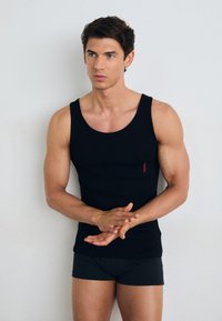 Schwarzes Tanktop aus glattem Stoff, mit rundem Ausschnitt und kleinem roten Logo-Akzent. Getragen mit passenden dunklen Boxershorts.