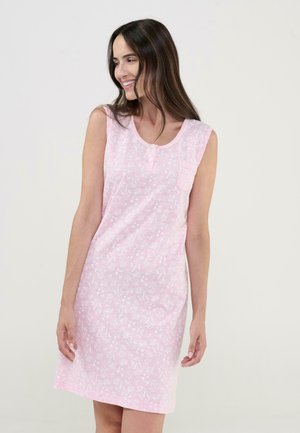 Mujer con vestido sin mangas de flores rosas hasta la rodilla, sonriendo y mirando hacia su izquierda sobre un fondo liso.