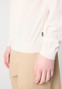 Pull en tricot rose clair avec des poignets et un ourlet côtelés. Présente une petite étiquette de marque noire sur le côté, porté sur un pantalon beige.