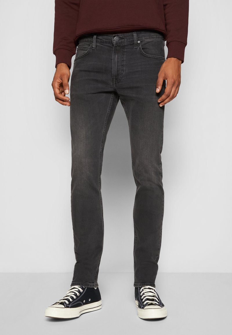 Homme portant un jean slim gris foncé, des baskets en toile noires et blanches, et un haut à manches longues bordeaux foncé, debout devant un fond clair.