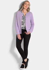 Lichtpaarse cardigan met ronde hals en knopen, gecombineerd met een zwart topje met patroon, zwarte skinny jeans en zwarte sneakers met witte zolen.