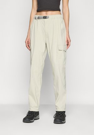 Personne portant un pantalon cargo beige avec des poches latérales et une ceinture noire, associé à des chaussures de randonnée noires et un top court noir.