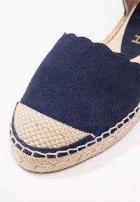 Gros plan d’une espadrille en daim bleu marine avec un bout en jute tissé et une couture tressée sur la semelle, sur fond blanc.