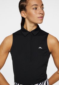 Svart ärmlös polo med knäppkrage och en liten vit logotyp. Slätt, stretchigt tyg med en figurnära design. Minimalistisk stil.