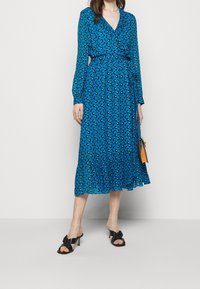 Vestido azul con estampado, de mangas largas, escote en V y cuello con volantes. Presenta una cintura fruncida y un dobladillo acampanado. Sandalias negras de tacón y bolso amarillo.