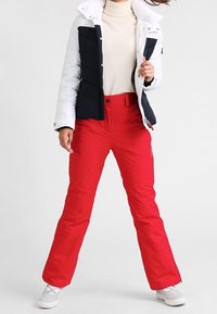 Pantalon de ski rouge à coupe droite, associé à une veste matelassée blanche et noire. La tenue comprend des manches longues et un col roulé.